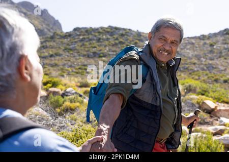 Felice coppia biraciale senior con zaini, mani in mano e trekking con bastoni da trekking Foto Stock