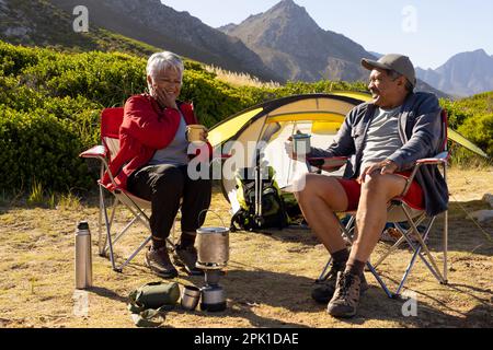 Felice coppia biraciale anziana seduta su sedie da campeggio e bere caffè Foto Stock