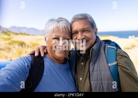 Felice coppia biraciale senior che indossa zaini, che prende selfie e sorridendo Foto Stock