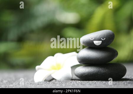 Pila di pietre con il volto felice disegnato e fiore su tavolo grigio all'aperto, spazio per il testo. Siate in armonia e godetevi la vostra vita Foto Stock