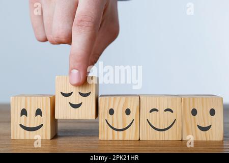 Uomo che organizza cubi con emoticon diverse in fila su un tavolo di legno su sfondo chiaro, primo piano Foto Stock