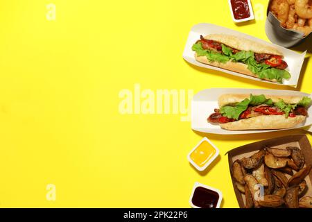 Gustosi hot dog, spicchi di patate, anelli di cipolla fritti e salse diverse su sfondo giallo, piatto con spazio per il testo. Fast food Foto Stock