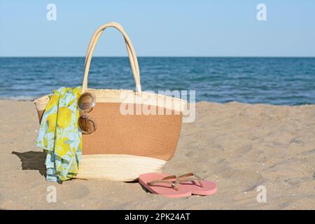 Borsa in paglia con impacco da spiaggia, occhiali da sole e infradito sulla spiaggia sabbiosa, spazio per testo. Accessori estivi Foto Stock