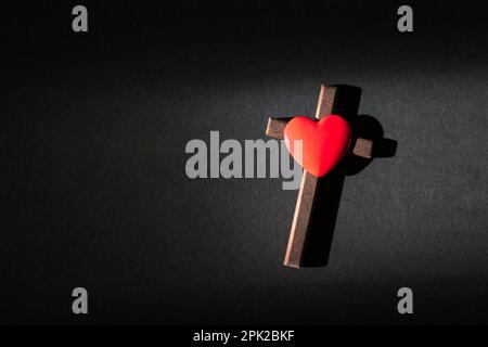 Cuore rosso in cima alla croce di legno crocifisso su sfondo nero. Vista dall'alto. Foto Stock