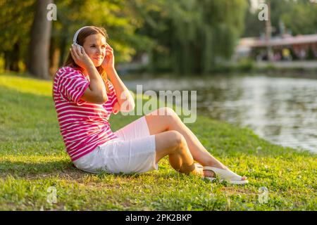 Donna rilassata seduta sull'erba ascoltando musica con le cuffie in un parco Foto Stock
