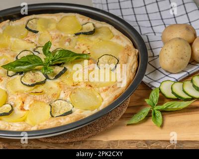 Una padella di quiche con patate e zucchine affettate su un tagliere di legno con patate intere accanto Foto Stock
