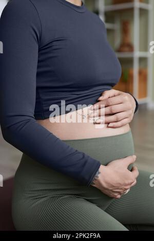 Primo piano verticale di una donna incinta che fa yoga prenatale con particolare attenzione alle mani che tengono il ventre tatuato Foto Stock