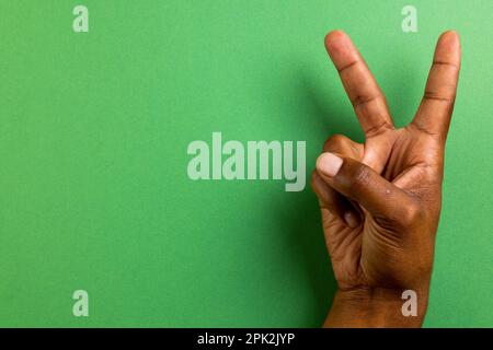 Primo piano di mano di un uomo biraciale che mostra un segno di pace con spazio copia su sfondo verde Foto Stock