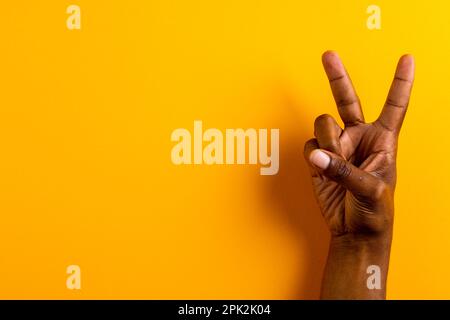 Primo piano di mano di un uomo biraciale che mostra un segno di pace con spazio copia su sfondo giallo Foto Stock