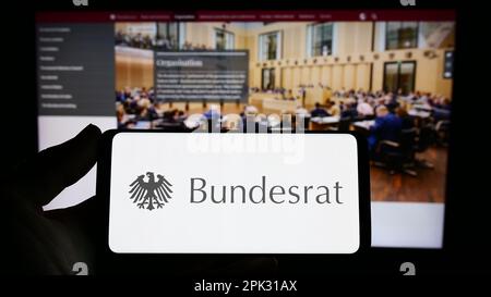 Persona che tiene in mano uno smartphone con il sigillo dell'organo legislativo tedesco Bundesrat sullo schermo di fronte al sito Web. Messa a fuoco sul display del telefono. Foto Stock