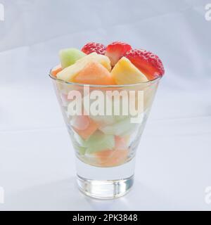 Macedonia di frutta fresca di fragole, meloni di melone e meloni di melone e ananas in una tazza di vetro trasparente su fondo di lino bianco Foto Stock