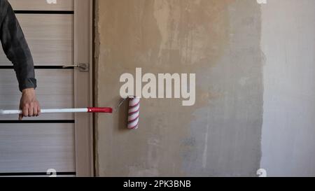 la parete della stanza in fase di applicazione del primer con un rullo striato su un lungo manico nelle mani del pittore Foto Stock