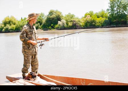 Uomo premuroso in camo vestito in piedi a poppa della barca, la pesca, mentre il cane riposa accanto fuori. Foto Stock