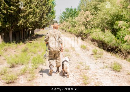Irriconoscibile maschio pescatore in camo vestito a piedi lungo strada sterrata con cane macchiato in estate. Foto Stock