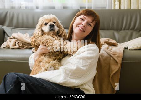Una giovane donna tiene in braccio un americano Cocker Spaniel mentre si siede sul pavimento vicino al divano nella stanza. Retroilluminazione dalla finestra. Foto Stock