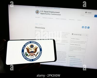 Persona che tiene un telefono cellulare con il logo della Commissione per le pari opportunità di lavoro (EOC) sullo schermo davanti alla pagina web. Messa a fuoco sul display del telefono. Foto Stock