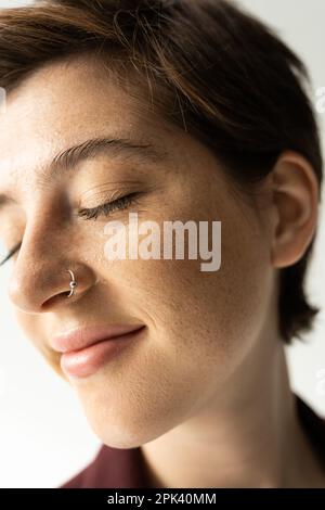 primo piano ritratto di donna spensierata con lentiggini e naso piercing sorridente con occhi chiusi su sfondo grigio, immagine stock Foto Stock