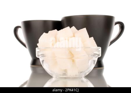 Qualche cubetto di zucchero bianco in una ciotola di vetro con due tazze nere, macro, isolata su fondo bianco. Foto Stock