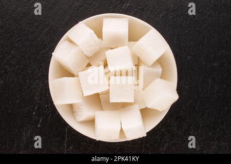 Diversi cubetti di zucchero bianco con piattino di ceramica bianco su pietra ardesia, macro, vista dall'alto. Foto Stock