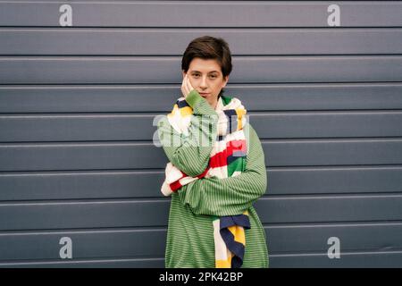 donna scettica in pullover verde e sciarpa multicolore a righe posa con mano vicino al viso a parete grigia sulla strada, immagine stock Foto Stock