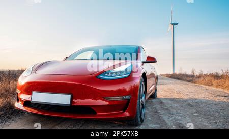 Vista di un'auto elettrica rossa parcheggiata. Turbina eolica sullo sfondo Foto Stock