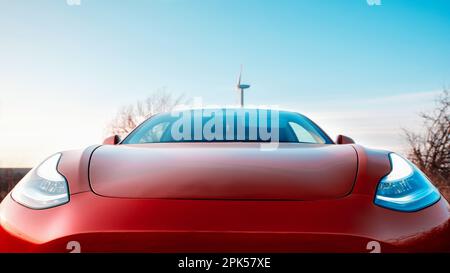 Vista frontale ravvicinata di un'auto elettrica rossa parcheggiata. Turbina eolica sullo sfondo Foto Stock