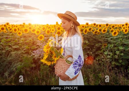 Giovane donna che cammina nel campo di girasole fiorente al tramonto tenendo sacchetto di paglia con fiori. Paesaggio estivo. Sentirsi in armonia con la natura Foto Stock