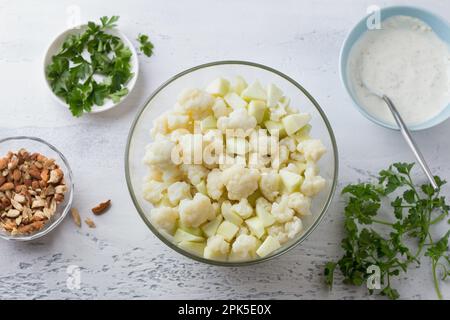 Ciotola di vetro con cavolfiore al vapore mescolato con mele verdi, circondata da ingredienti per insalata: Mandorle, condimento allo zenzero yogurt e prezzemolo , vista dall'alto. Foto Stock