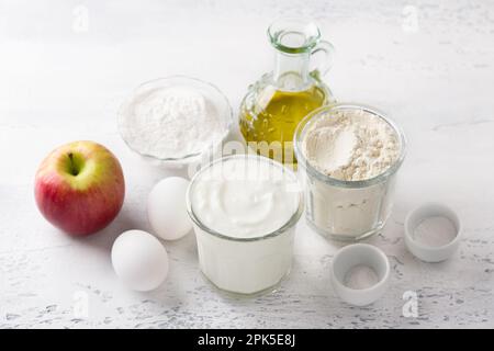 Ingredienti per frittelle di mele: mela, yogurt, farina di grano e riso, uova, olio vegetale, sale e lievito in polvere su sfondo grigio chiaro, vista dall'alto. Foto Stock