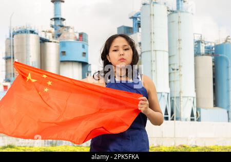 Preoccupata ragazza asiatica in abiti da lavoro con la bandiera della cina in piedi di fronte a scenario industriale Foto Stock