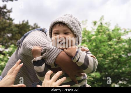 Primo piano del nuovo papà che gioca con il bambino, sollevando, gettando il bambino cute in su nell'aria nella natura. Ragazzo sorridente che tiene con forza le braccia del padre, le mani della madre. M Foto Stock