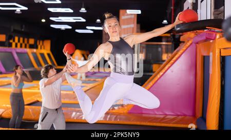 Donna di mezza età che lancia la palla nel basket alto saltando in trampolino arena Foto Stock