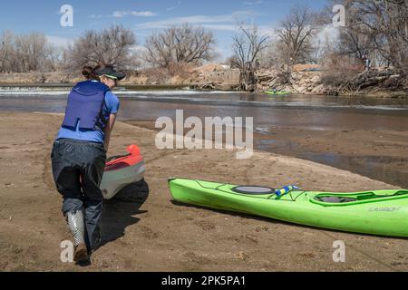 Evans, CO, USA - 1 aprile 2023: I paddlers stanno portando i kayak sopra una diga di diversione durante il viaggio primaverile in canoa sul fiume di South Platte a Colora Foto Stock