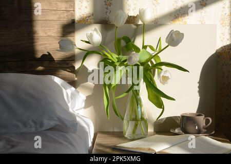 Bel bouquet di tulipano bianco, tazza di bevanda e libro aperto sul comodino in camera da letto Foto Stock