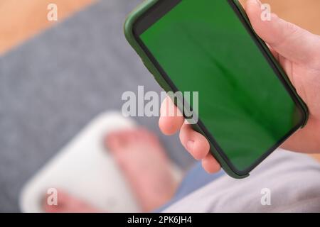 Un uomo si trova sulla bilancia. Il ragazzo sta pesando se stesso. Bilance da pavimento per la salute. Controllo del peso. Schermo verde dello smartphone. Foto Stock