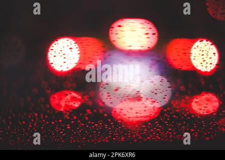 Luci bokeh rosse lucide che si illuminano dietro la finestra in vetro bagnato con gocce di pioggia nelle ore notturne scure Foto Stock