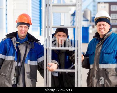 Tre lavoratori in tute invernali e caschi stanno fuori guardando la fotocamera e sorridendo. Vero ritratto di operai caucasici. Ci sono dei costruttori felici con una scala portatile all'esterno. Foto Stock