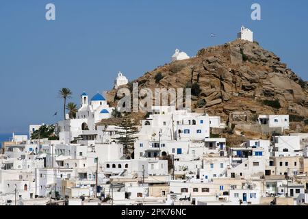 Vista panoramica di una chiesa, cappelle più piccole e la bandiera greca sulla cima di una collina che domina il Mar Egeo a iOS in Grecia, noto anche come Chora Foto Stock