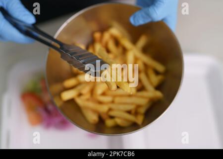 Il cuoco tiene una ciotola con patatine fritte. L'addetto alla cucina ha messo i bastoncini di patate fritti in una scatola per il pranzo per portarli via Foto Stock