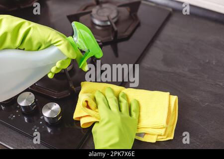 La mano della donna in guanto verde spruzza detergente ecologico a fornello a gas. Primo piano della stufa in vetro Foto Stock