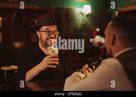 l'uomo si diverte a chiacchierare con un barista al bancone del bar mentre beve birra da un bicchiere in un pub Foto Stock