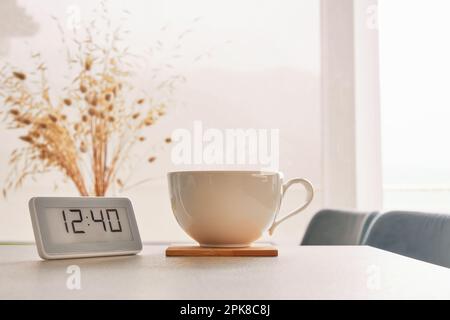 Concetto di tempo di pausa caffè. Una tazza di caffè caldo e un orologio digitale sul tavolo. Interni accoglienti sfondo a tema Foto Stock
