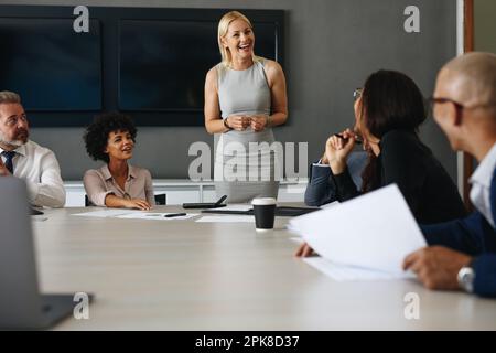 Una donna felice che sorride mentre si alza e parla con il suo team in una riunione. Imprenditrice femminile che prende il comando in una discussione aziendale, Foto Stock