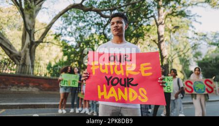 Giovane attivista che guarda via mentre tiene un cartello durante un raduno sul cambiamento climatico. Ragazzo adolescente protestante contro il riscaldamento globale con un gruppo di d Foto Stock