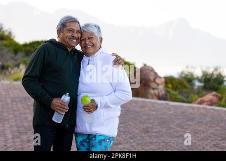 Ritratto di felice coppia biraciale anziana che indossa abbigliamento sportivo, abbracciando in strada in montagna Foto Stock