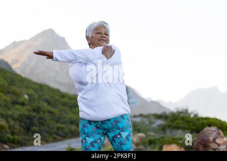 Felice donna biraciale anziana che indossa abbigliamento sportivo, che si allunga per strada in montagna Foto Stock