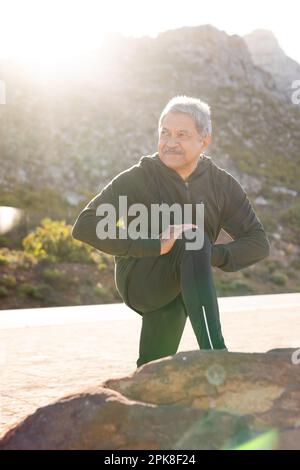 Felice uomo biraciale anziano che indossa abbigliamento sportivo, che si allunga per strada in montagna Foto Stock