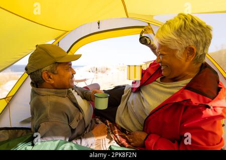 Felice coppia biraciale anziana sdraiata in tenda e bevendo caffè in montagna Foto Stock