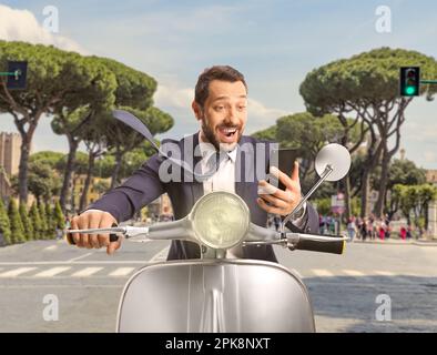 Felice giovane uomo in sella a uno scooter per le strade di Roma e guardando uno smartphone Foto Stock