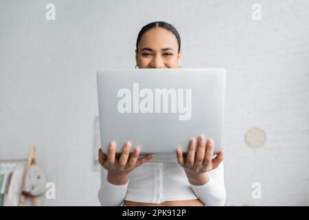 felice studente afro-americano che copre metà del volto mentre tiene il portatile, immagine di borsa Foto Stock